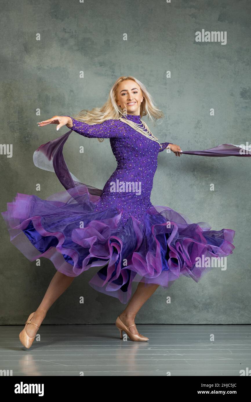 Ballerina Bionda femmina in movimento di danza scorrevole in abito da ballo viola, sullo sfondo grigio dello studio fotografico Foto Stock