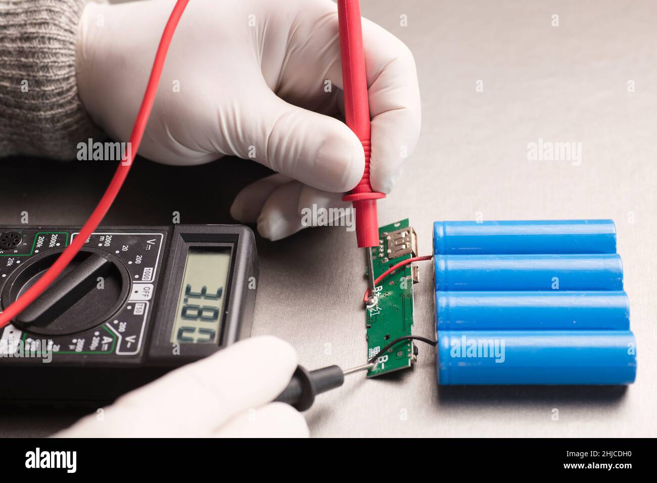 Circuito indicatore di carica, multimetro con poli rosso e nero nelle mani di uomo che controlla la tensione e gli ampere, 18650 batterie blu, sfondo grigio. Powerba Foto Stock