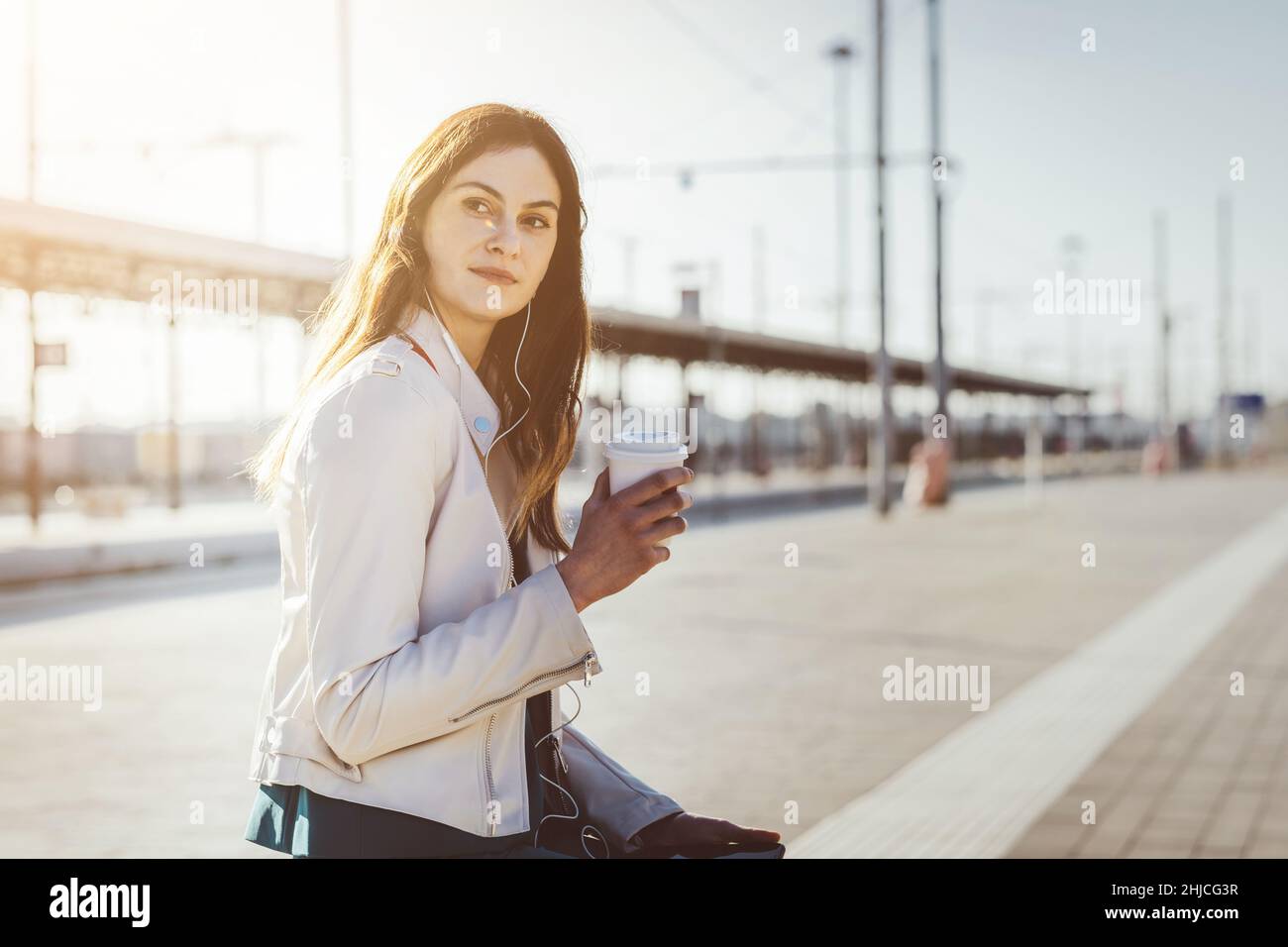 Una bella giovane donna in stazione sorridente e in attesa del treno al binario seduto sul bagaglio e con smartphone Foto Stock