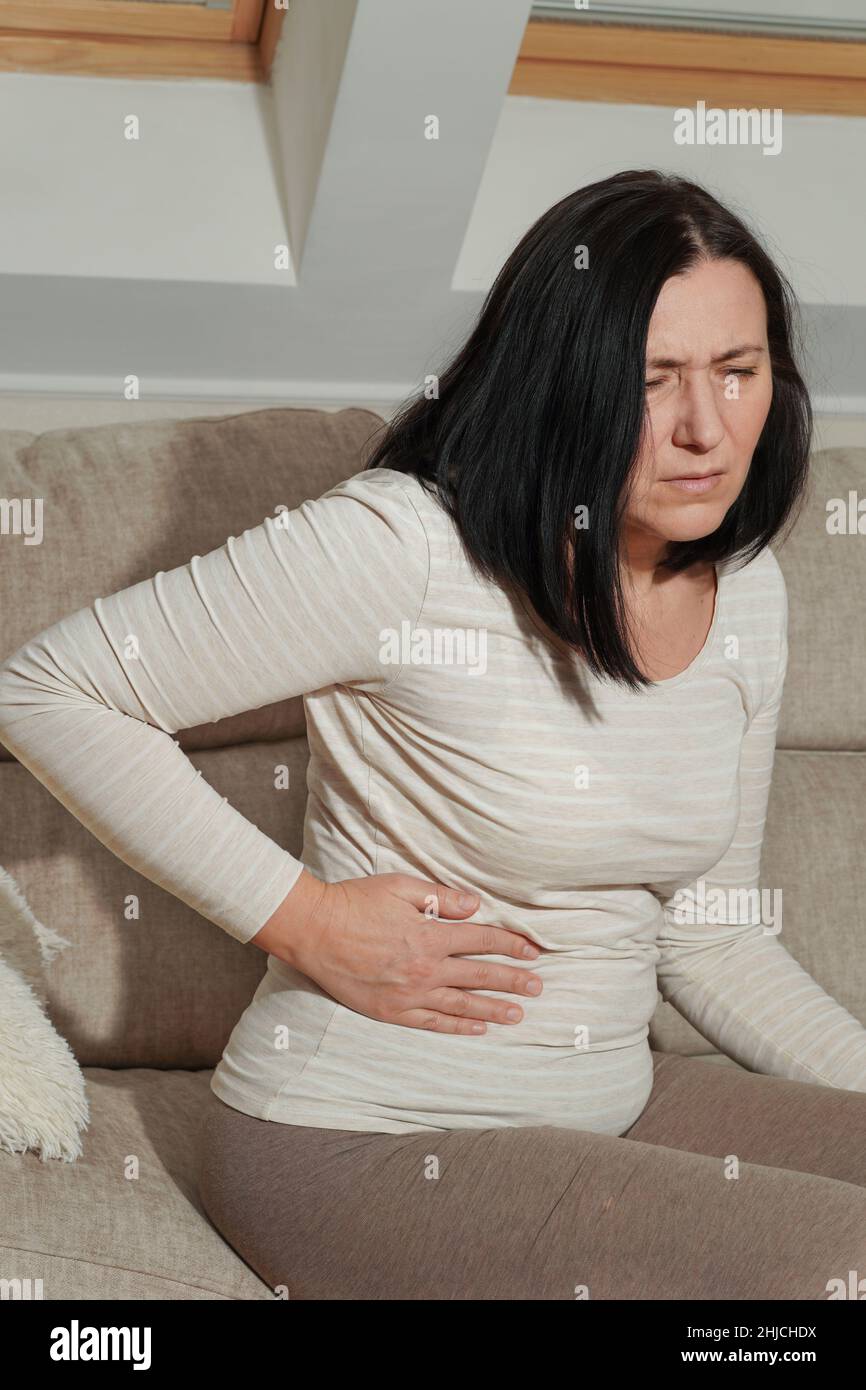 Donna matura di mezza età che soffre di mal di schiena e dolore al fianco a casa. Malattia, ilness, tristezza e turbato. Sintomi di disperazione o di vita malsana Foto Stock