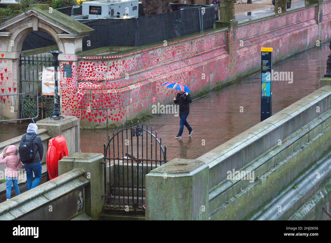 Un pedone che tiene un ombrello con bandiera dell'Unione passa davanti ai cuori rossi sul National Covid Memorial Wall nel centro di Londra. Foto Stock