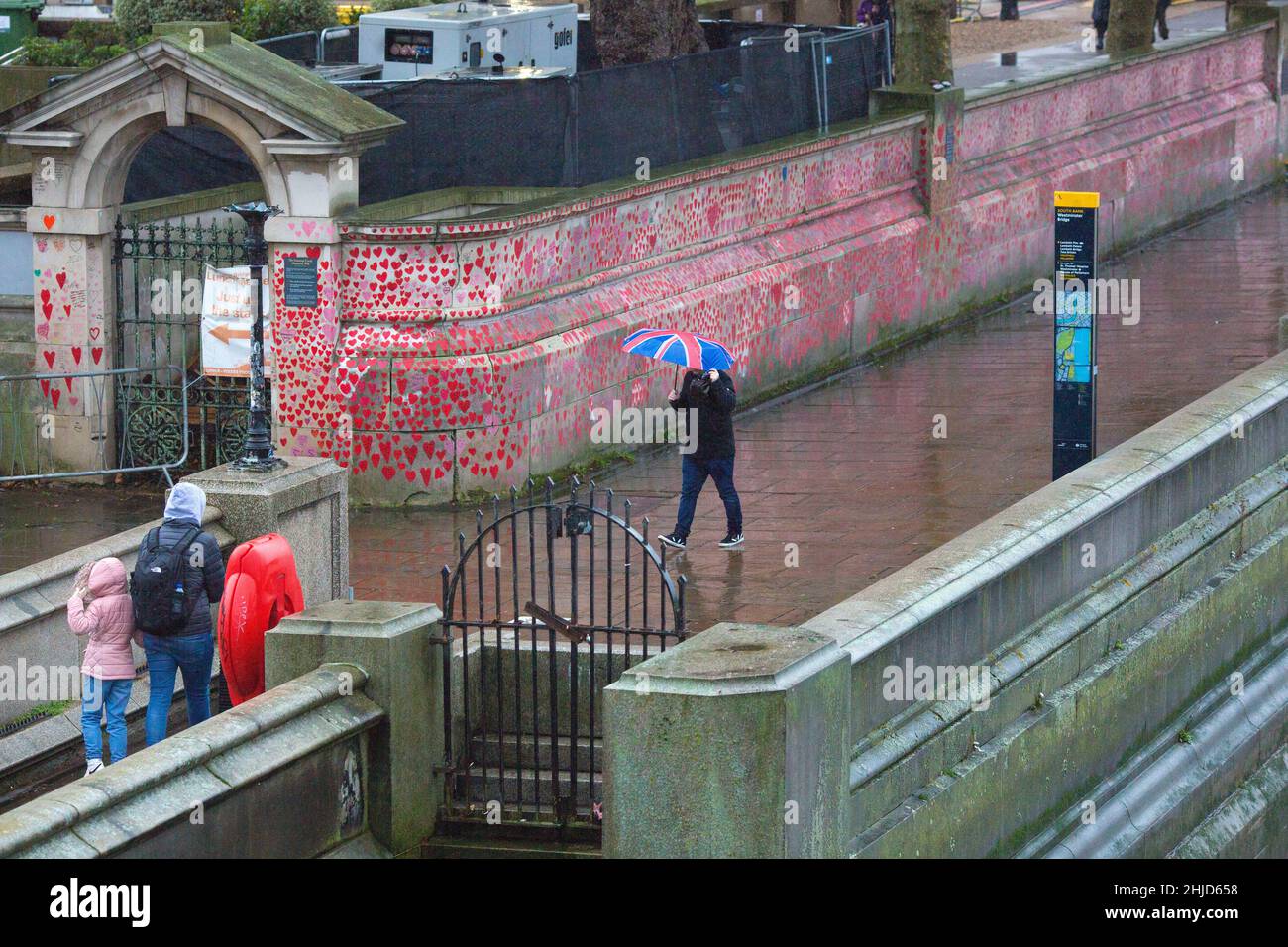 Un pedone che tiene un ombrello con bandiera dell'Unione passa davanti ai cuori rossi sul National Covid Memorial Wall nel centro di Londra. Foto Stock
