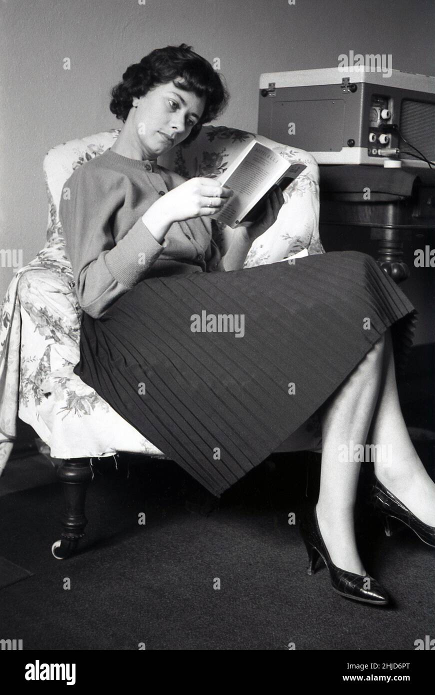 1950s, storica, all'interno di una stanza, seduta su una sedia, una giovane donna elegantemente vestita che indossa una blusa di lana e una gonna pieghettata, leggendo un libro cartaceo sulla pittrice italiana Botticelli, con un discografo portatile su un tavolo accanto a lei, Inghilterra, Regno Unito Foto Stock