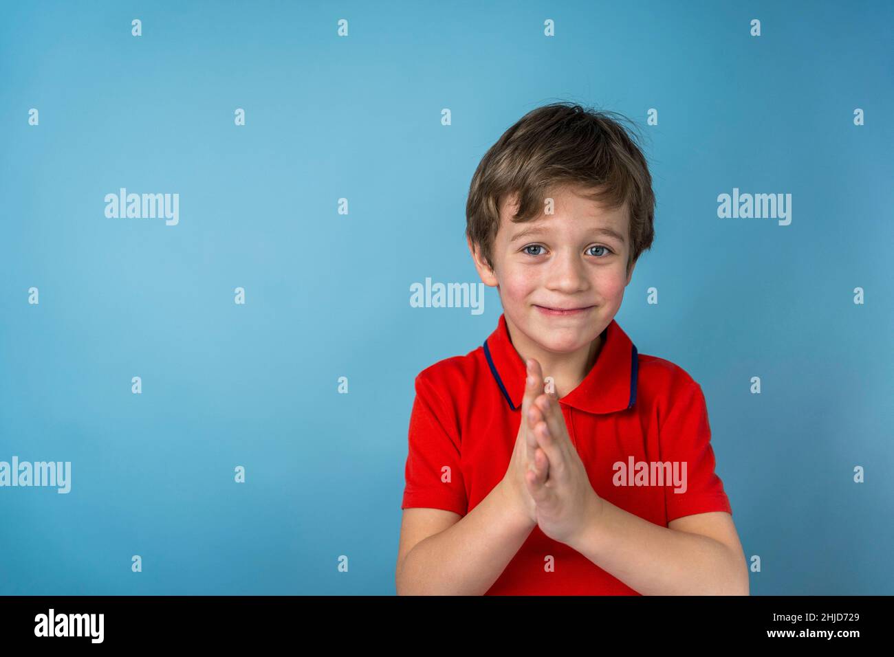 Un simpatico ragazzo caucasico di 5 anni in una T-shirt rossa si sfrega mano in mano e sorride slyly. Il ragazzo è contento del risultato, un'emozione positiva. Retro blu Foto Stock
