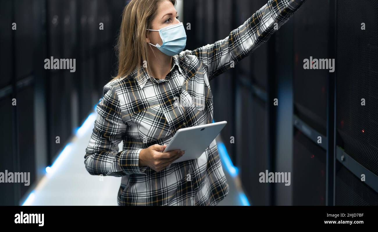 Tecnico del data center femminile che lavora all'interno della sala rack del server indossando la maschera facciale Foto Stock