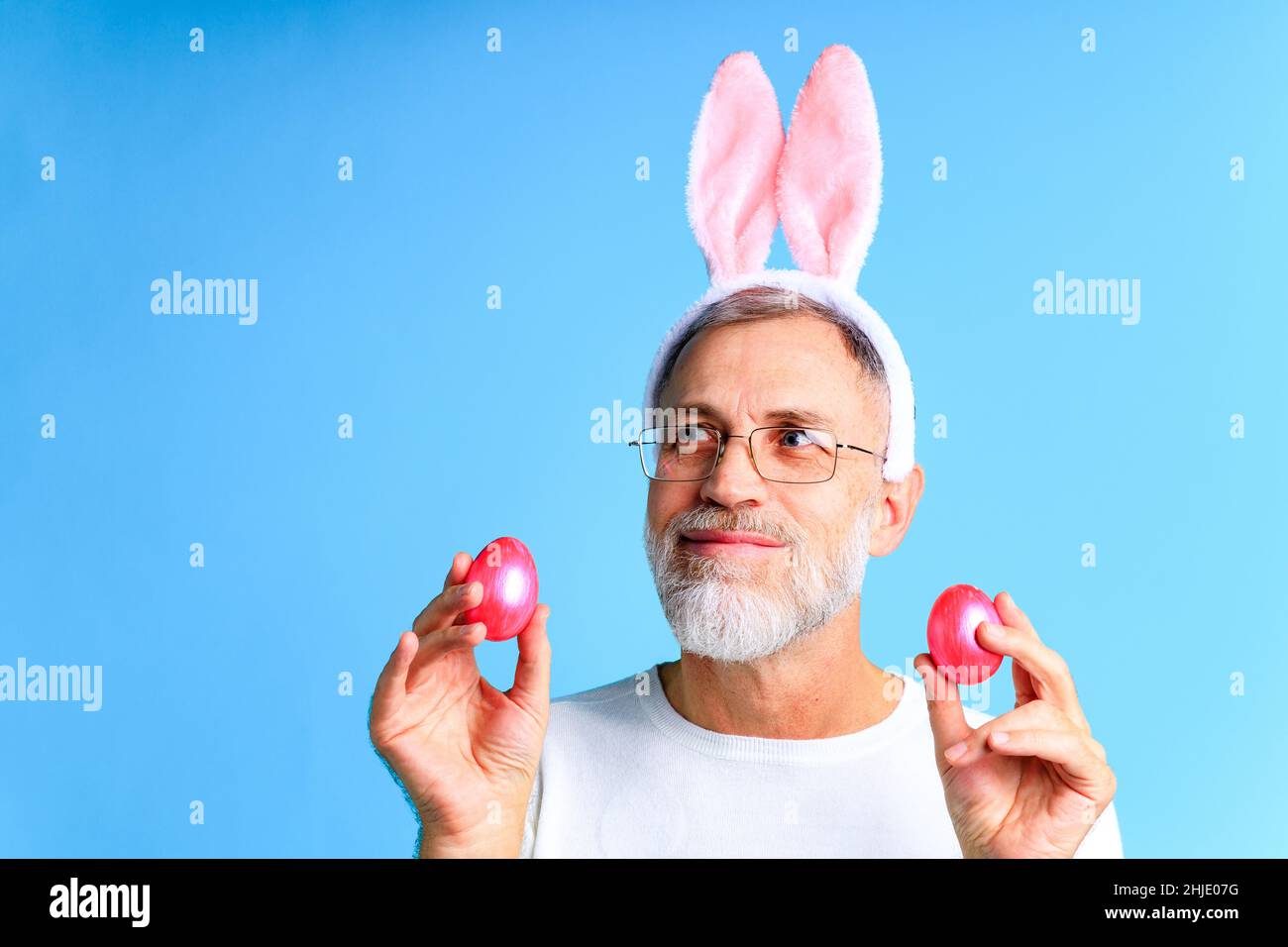 Carino uomo maturo con orecchie conigliate che tengono l'uovo di Pasqua su sfondo di colore blu Foto Stock