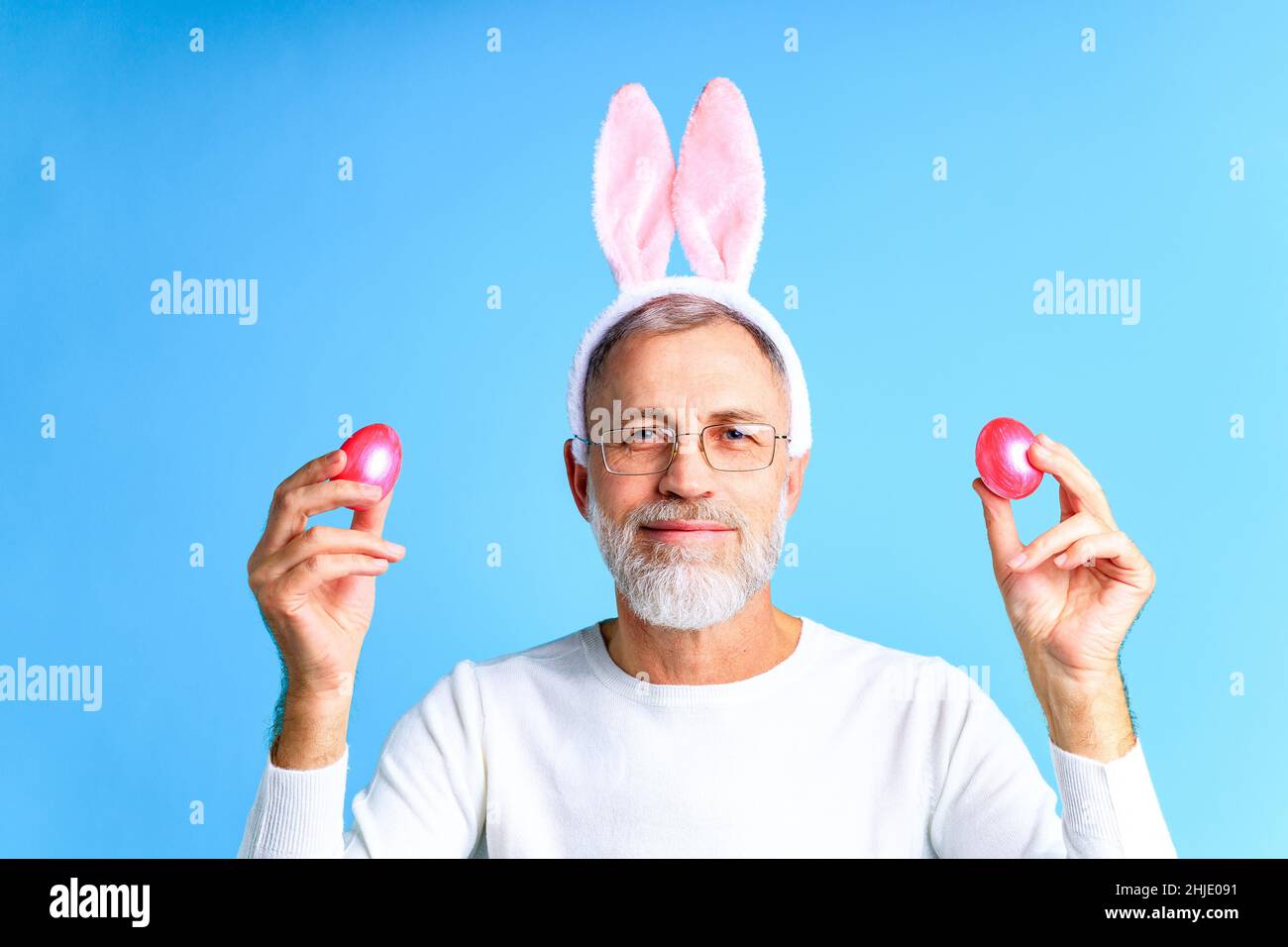 Carino uomo maturo con orecchie conigliate che tengono l'uovo di Pasqua su sfondo di colore blu Foto Stock