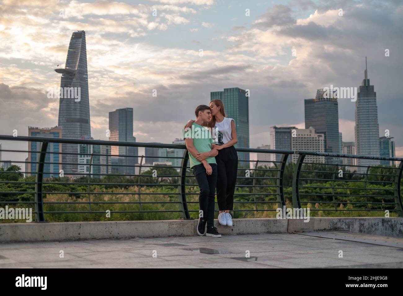 Bella coppia mista in piedi sul ponte nel parco in metropoli e coccole. La ragazza bacia il ragazzo. Grattacieli sullo sfondo Foto Stock