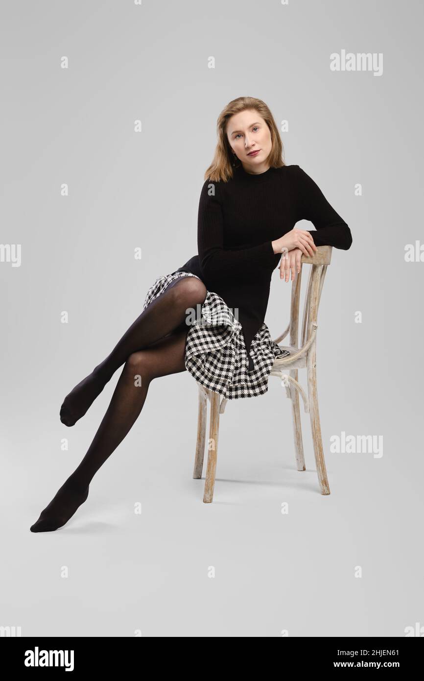 Donna a piedi nudi in dolcevita e gonna seduta su sedia in studio su sfondo grigio Foto Stock