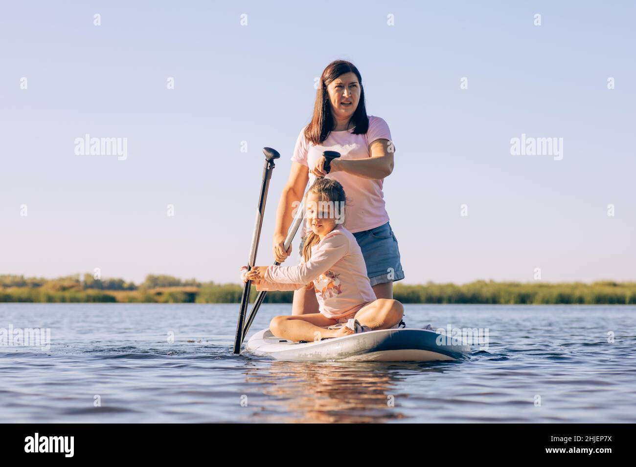 Donna di mezza età e figlia canottaggio seduta su bordo di sup con grandi sforzi, esercitandosi e godendo il tempo con canne verdi e alberi in background Foto Stock