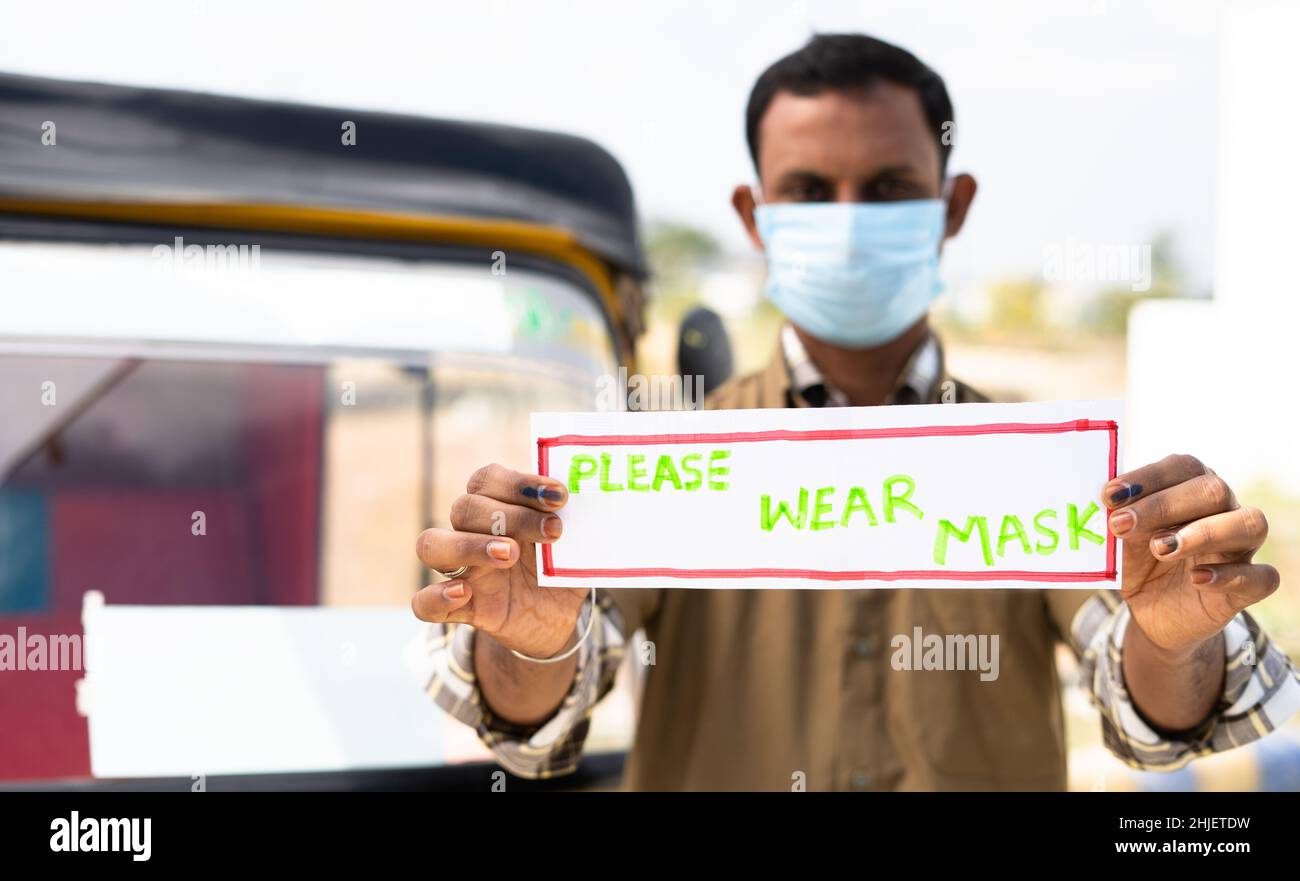 Focus a portata di mano, auto risciò conducente con maschera medica mostrando si prega di indossare maschera segno guardando la telecamera per il passeggero da proteggere Foto Stock