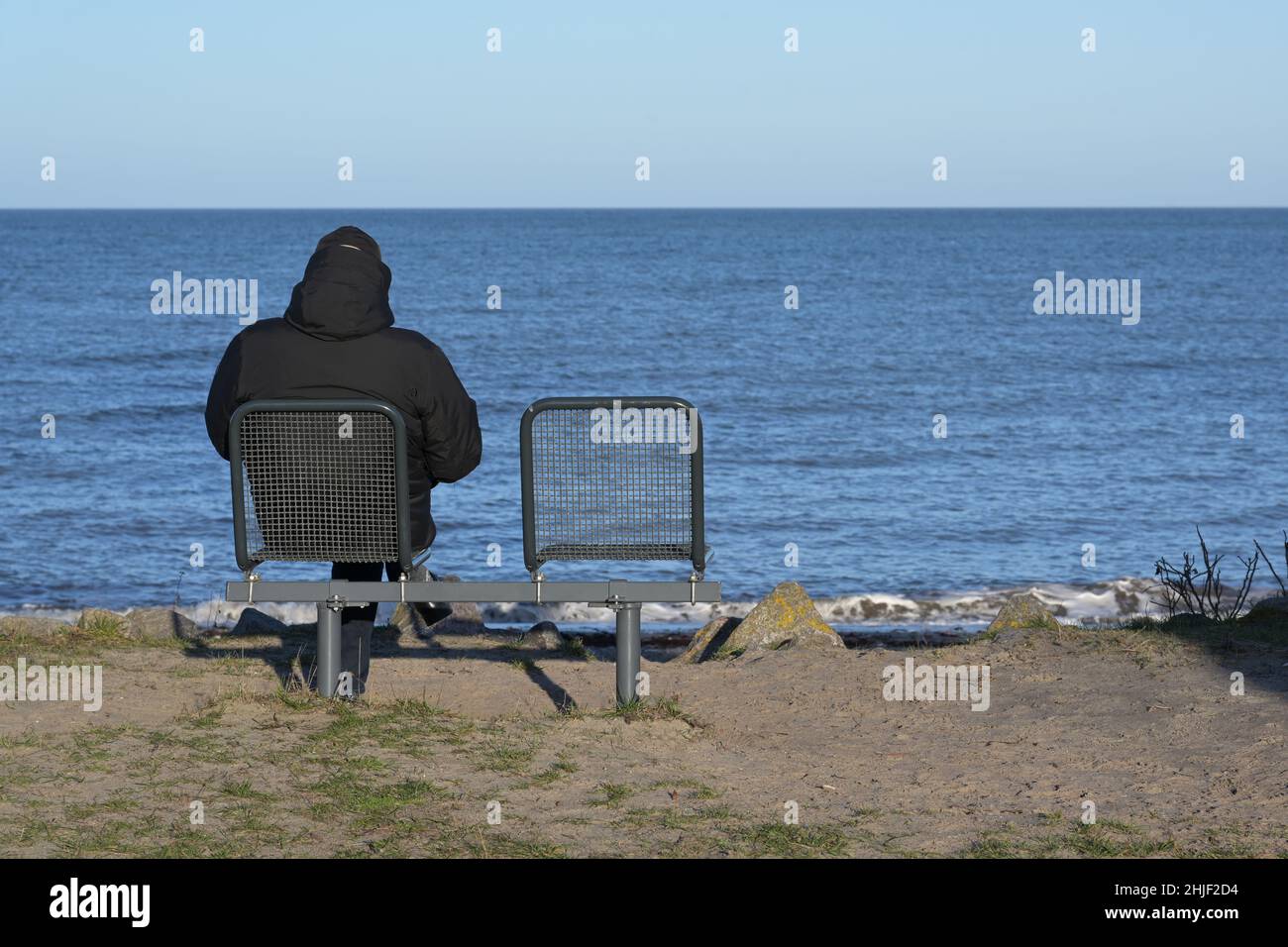Uomo singolo in una giacca calda e scura da dietro seduto accanto a una sedia vuota sulla riva del mare che guarda sull'acqua blu fino all'orizzonte, concetto per r Foto Stock