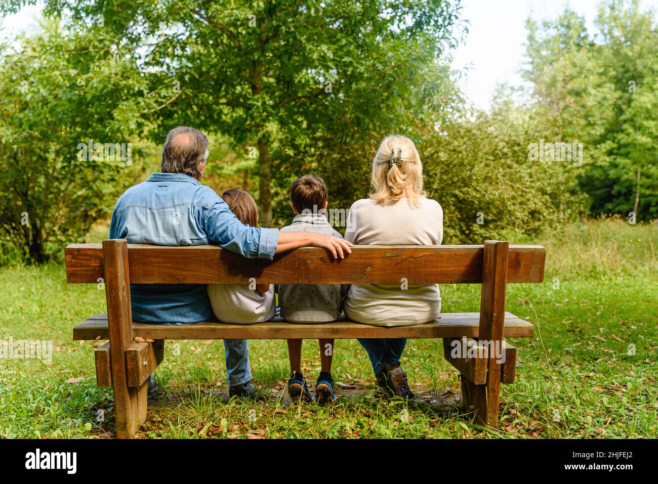 I nipoti e i loro nonni seduti insieme su una panchina nel parco circondato da alberi Foto Stock