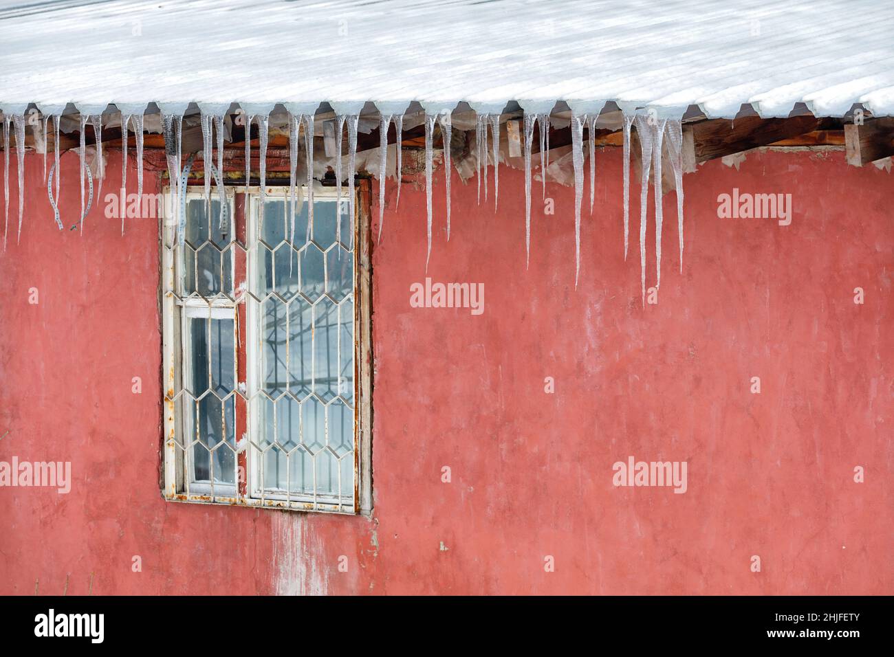 Ghiaccioli appendono dal tetto sopra la finestra barred di una vecchia casa con un muro rosso. Foto Stock