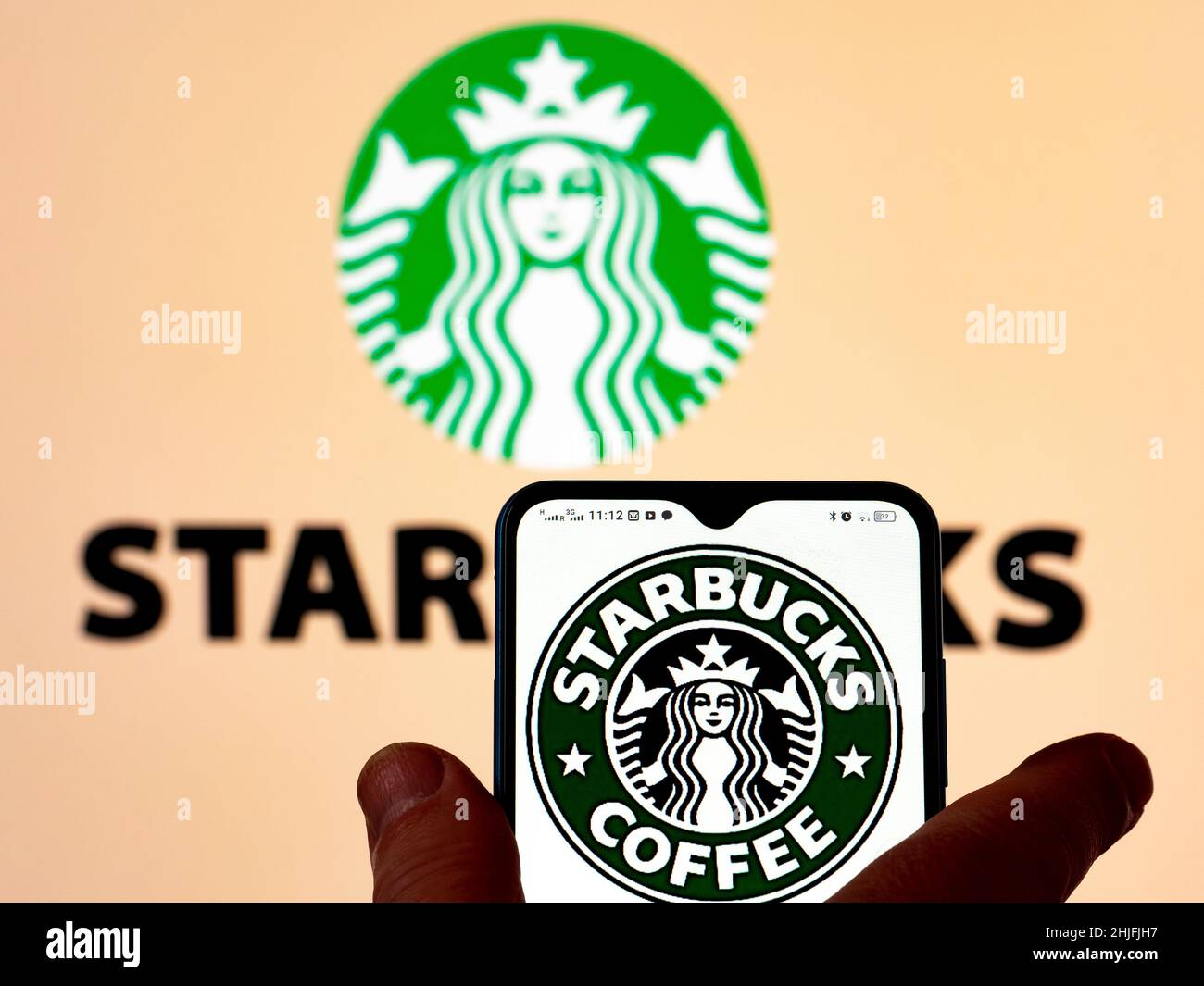 In questa figura, il logo del caffè Starbucks viene visualizzato sullo schermo di uno smartphone con il logo Starbucks Corporation sullo sfondo. Foto Stock