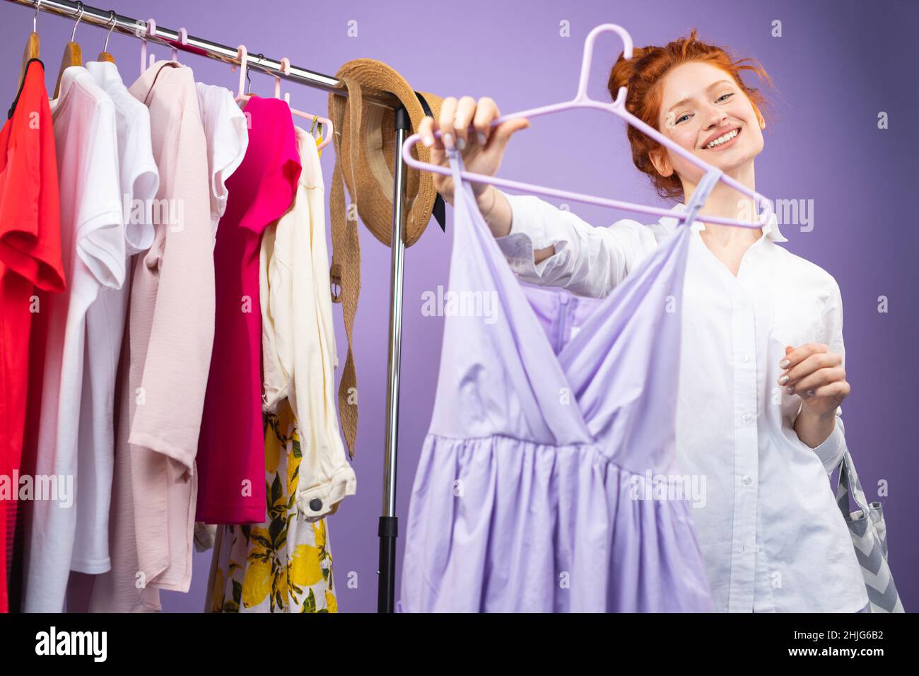 Una bella ragazza con capelli rossi accanto ad un vestito mostra un abito viola che ha scelto accanto ad una parete dello stesso colore Foto Stock