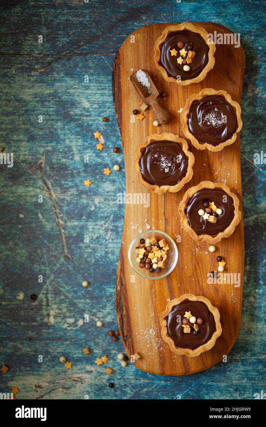 Cinque torte di caramello al cioccolato appena decorate su un asse di legno con una pentola di spruzzi accanto Foto Stock