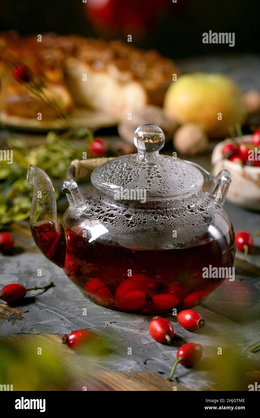 Frutti di bosco di rose tè alle erbe in teiera di vetro in piedi su vecchio tavolo in legno con frutti di bosco autunno e torta di mele intorno. Bevanda vitaminica calda. Foto Stock