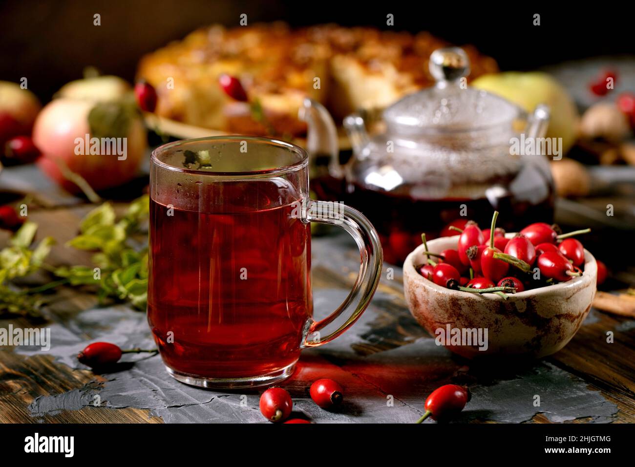 Rose hip bacche tè alle erbe in tazza di vetro in piedi su vecchio tavolo in legno con bacche selvatiche autunno e torta di mele intorno. Bevanda calda sana. rus scuro Foto Stock