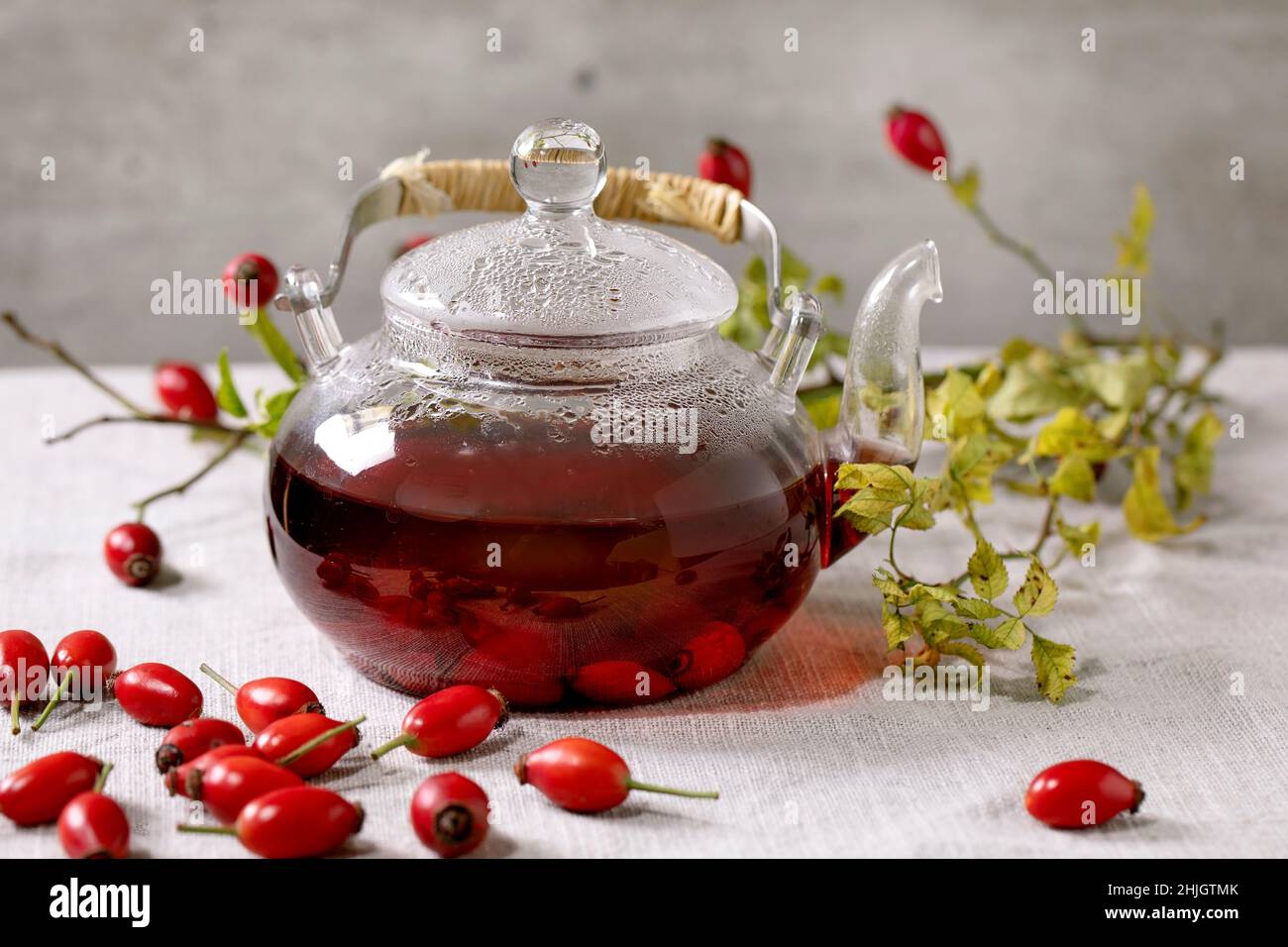 Rosa anca bacche tè alle erbe in teiera di vetro in piedi su panno da tavola di lino bianco con frutti di bosco autunno intorno. Bevanda calda sana. Foto Stock