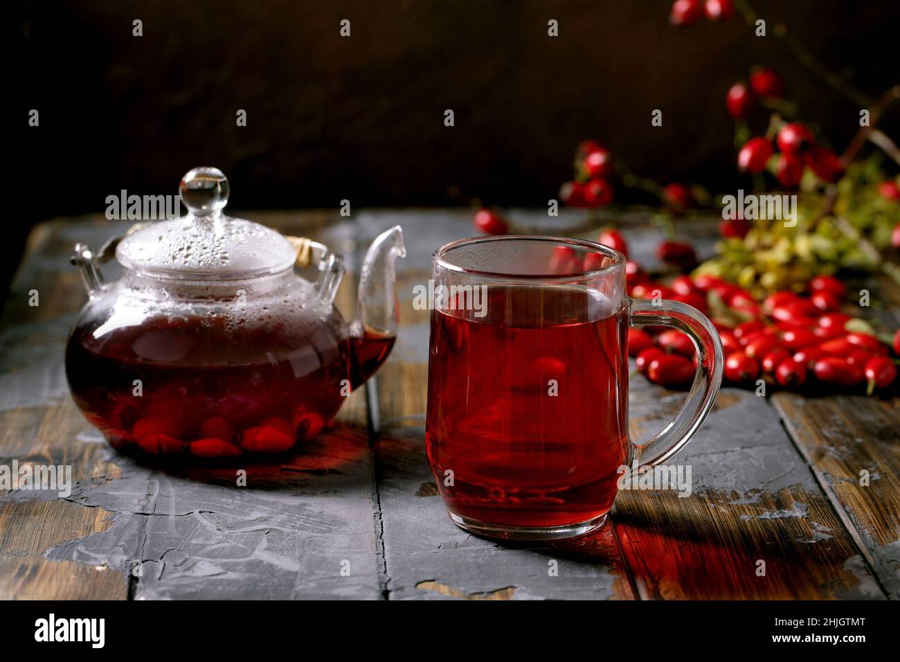 Tazza di frutti di bosco di rose e fianchi tè alle erbe e teiera di vetro in piedi su un vecchio tavolo in legno con frutti di bosco autunno intorno. Bevanda calda invernale. Foto Stock