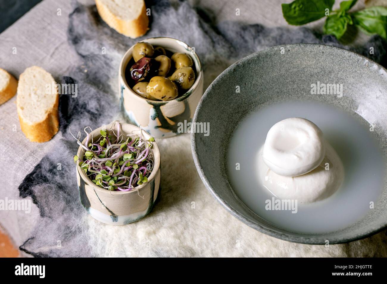 Burrata tradizionale italiana annodata formaggio in ceramica grigia ciotola sul tavolo. Pane, olive, insalata verde intorno. Ingredienti per un sano din mediterraneo Foto Stock