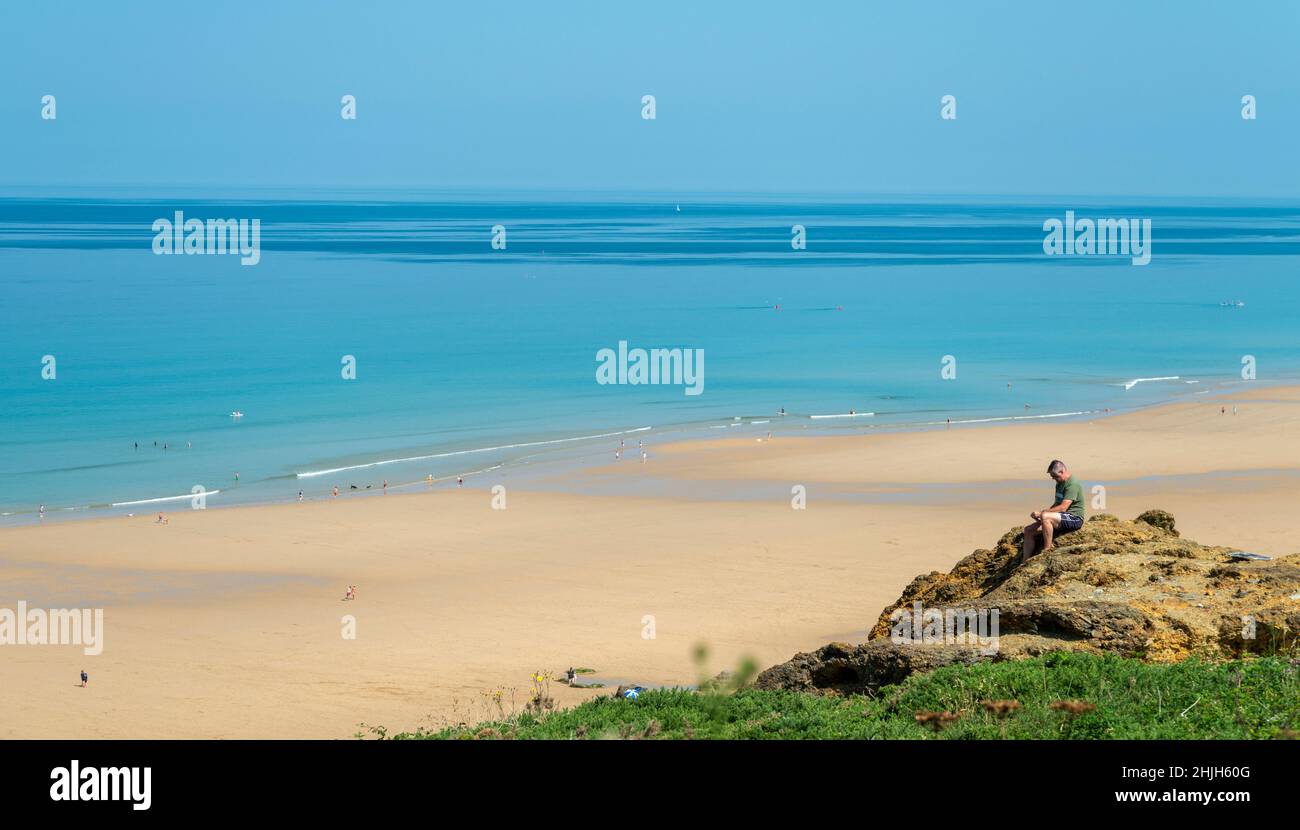Newquay,Cornovaglia,Inghilterra,Regno Unito-Giugno 21st 2021: Un uomo usa il suo telefono mentre si siede su una roccia nella calda metà estate Sunshine.overlooking the wi Foto Stock