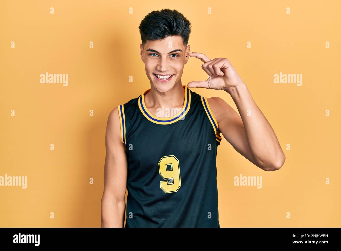 Giovane uomo ispanico che indossa uniforme basket sorridente e sicuro gesturing con mano che fa piccolo segno con le dita guardando e la fotocamera. mea Foto Stock