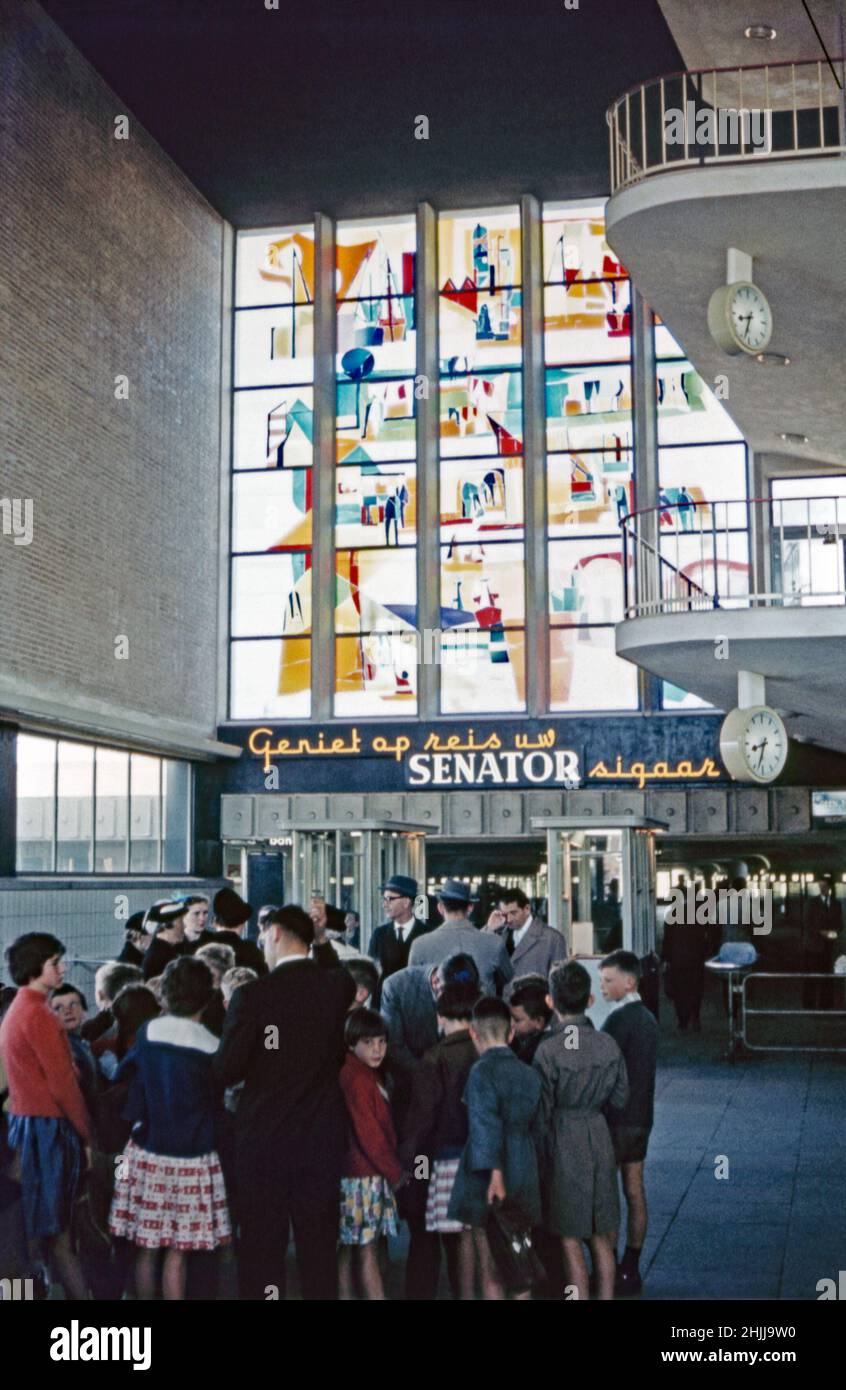 Passeggeri in attesa alla stazione ferroviaria Eindhoven Centraal, Eindhoven, Brabante Nord, Paesi Bassi c.1960. L'arte vetrata (vetrata applicata) è stata realizzata dall'artista olandese Lex Horn (1916-1968) e installata nella stazione nel 1955. Un cartello al neon pubblicizza i sigari senatori. La stazione ferroviaria centrale di Eindhoven è la stazione principale della città. Questa immagine proviene da una vecchia trasparenza del colore amatoriale in condizioni di scarsa illuminazione – una fotografia d'epoca 1950s/1960s. Foto Stock