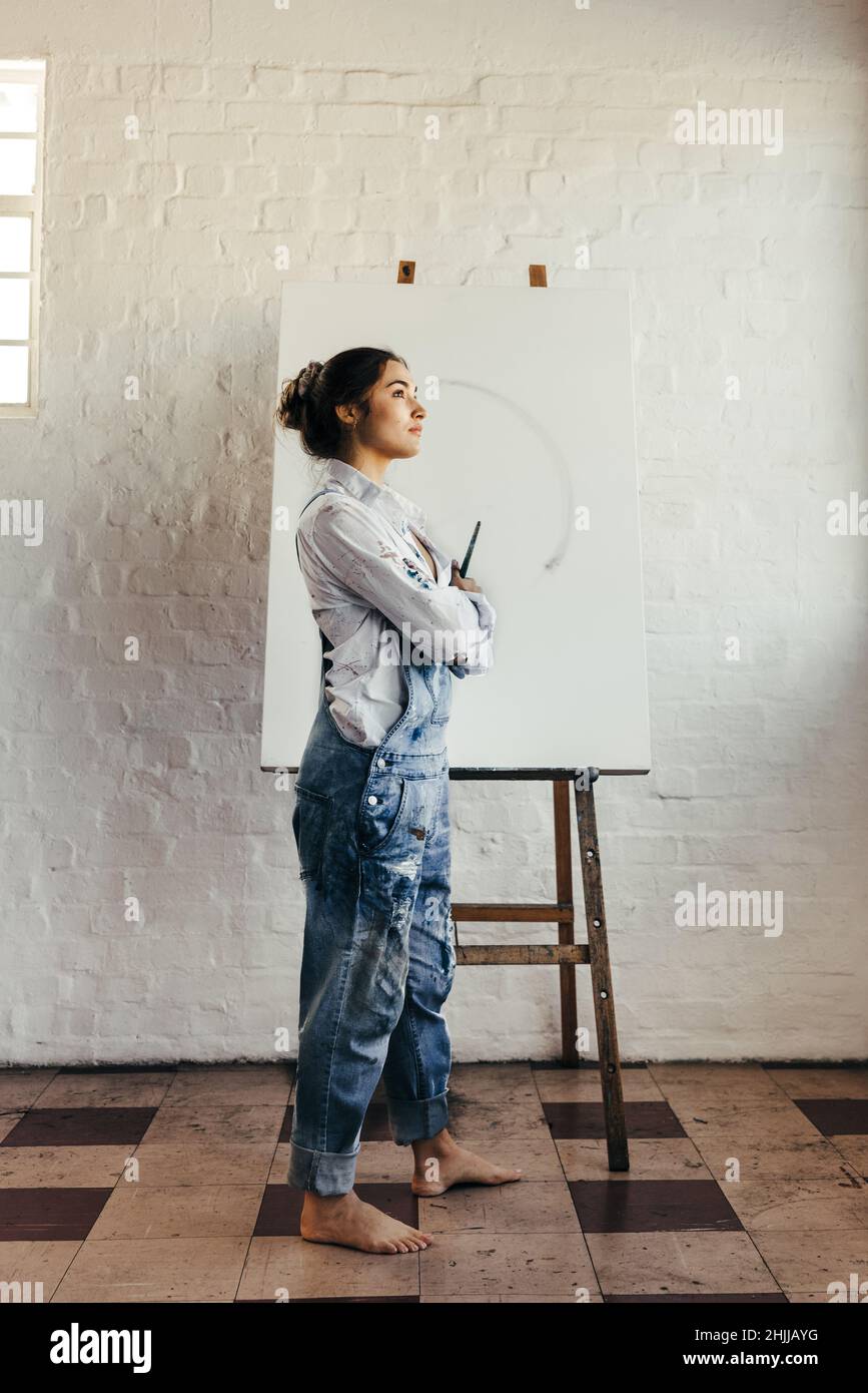 Artista femminile che pensa a nuove idee creative nel suo studio. Giovane pittore pensivo che guarda via mentre si alza di fronte a una tela bianca. Fantasioso Foto Stock