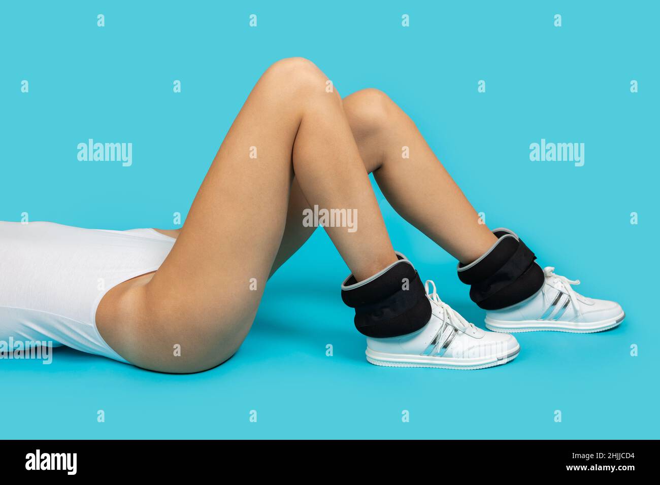 Adatta la giovane donna con le gambe lunghe indossando il bodysuit e le sneakers con pesi alla caviglia su sfondo blu facendo esercizi fisici. Stile di vita sano Foto Stock