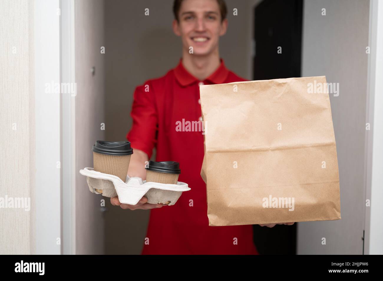 Uomo di deliveryman sorridente con cibo e caffè, ritratto di corriere in uniforme rossa con ordine, cibo da asporto ristorante, pacchetto ecologico e bicchieri kraft Foto Stock