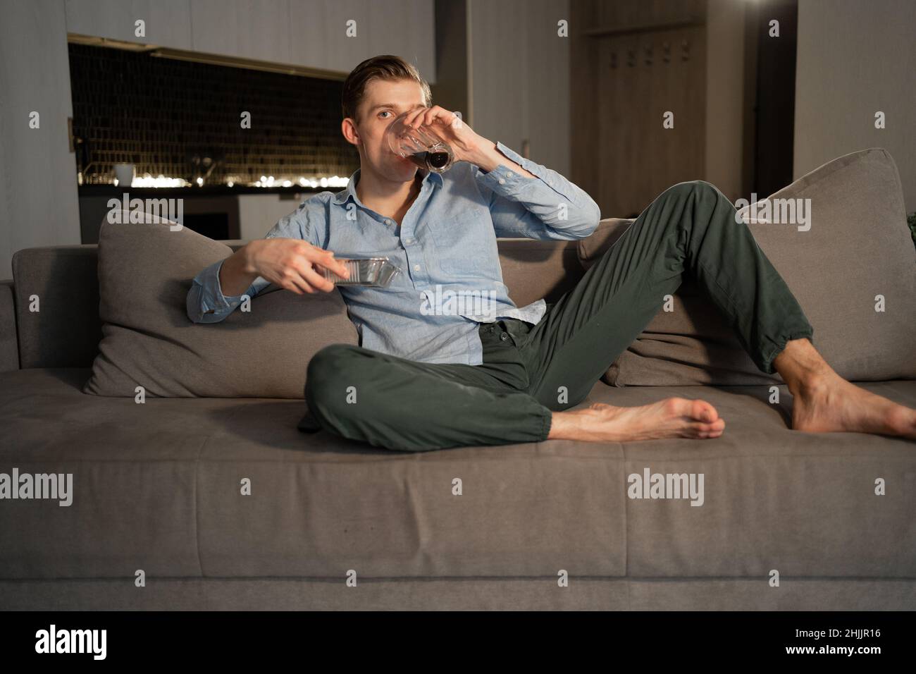 Un giovane guarda la TV a casa e beve una cola, rilassandosi e godendo la partita. Ragazzo caucasico sul divano che trascorre un fine settimana a casa Foto Stock