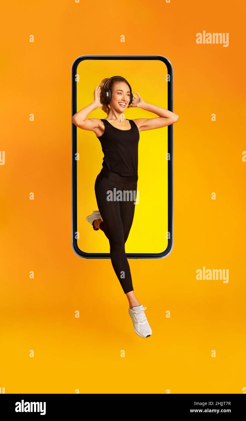 Sport Lady saltando dallo schermo del telefono indossando cuffie, sfondo giallo Foto Stock