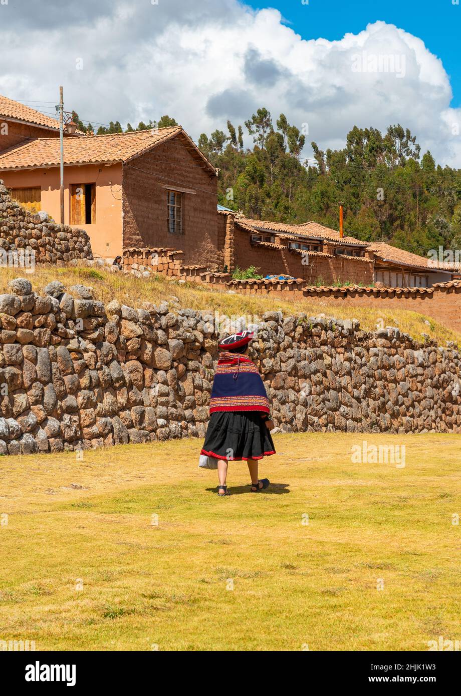 Donna indigena peruviana quechua in abbigliamento tradizionale che cammina di fronte a un muro inca della rovina Chinchero inca, Perù. Foto Stock
