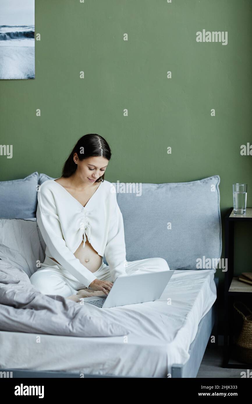 Giovane donna incinta che lavora online su un computer portatile mentre si siede sul letto in camera da letto Foto Stock