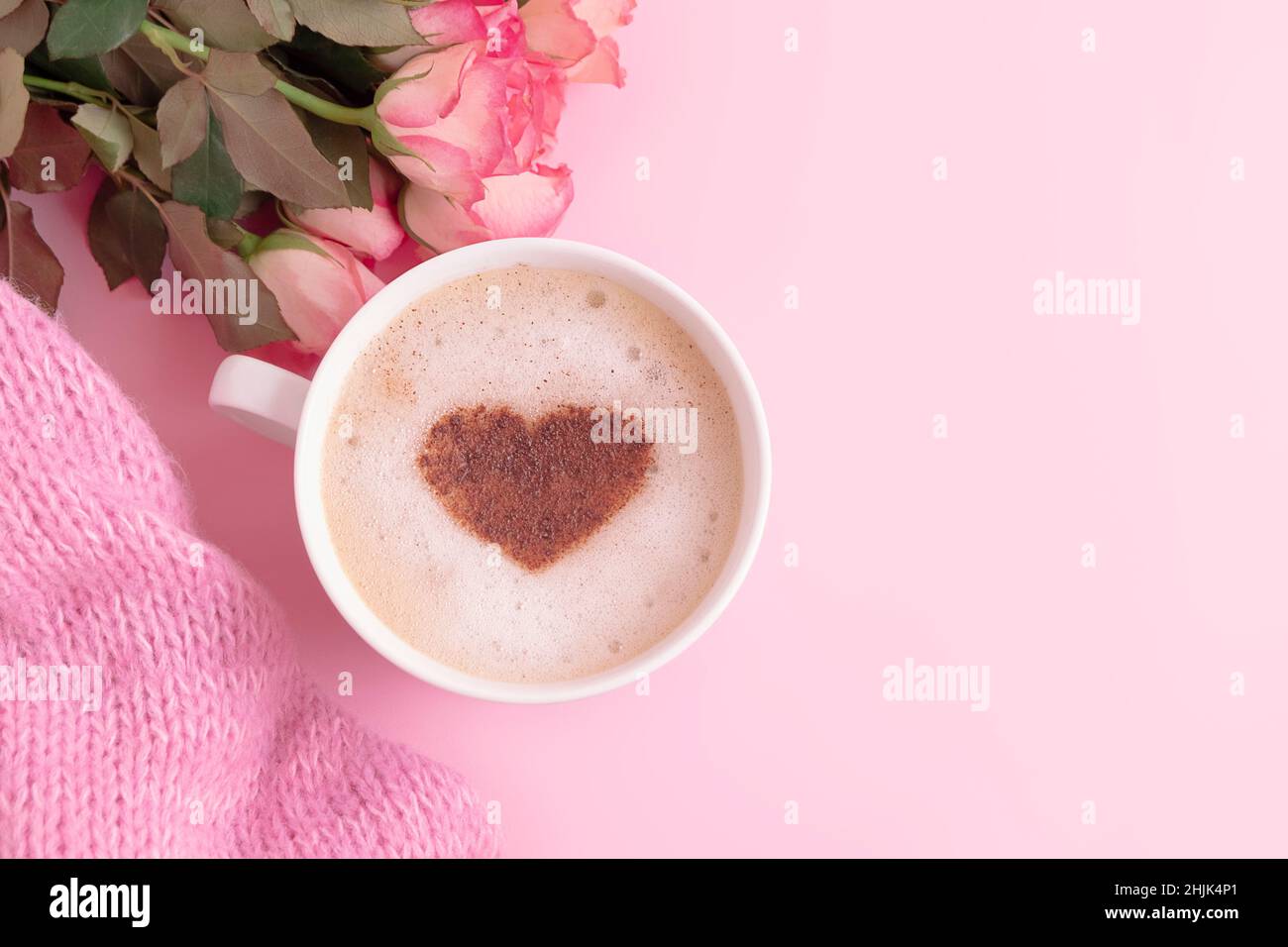 Composizione romantica con tazza di caffè con cuore sulla parte superiore, fiori e coperta in maglia su sfondo rosa pastello e spazio copia Foto Stock