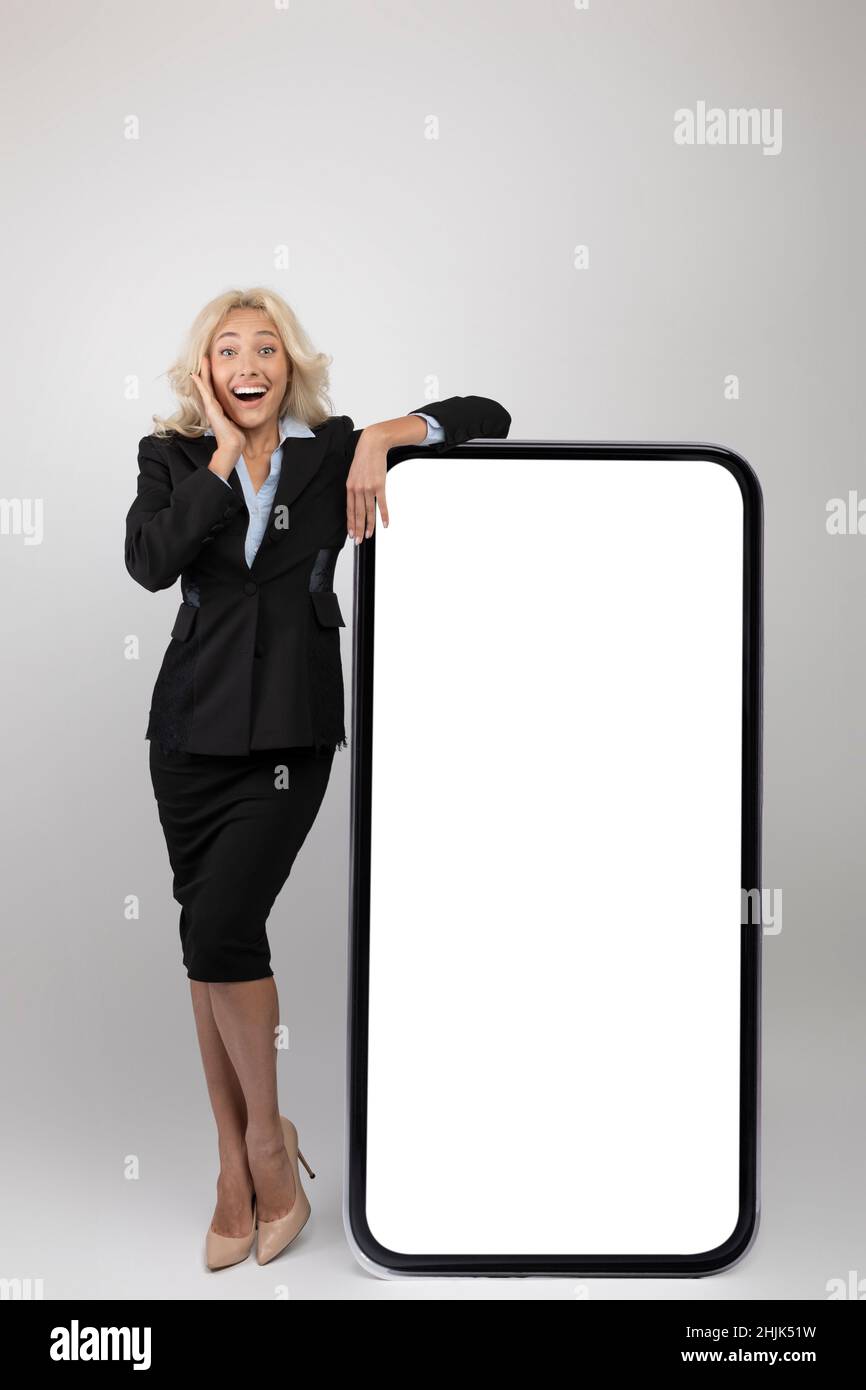 App aziendale. Entusiasta imprenditrice femminile in abbigliamento formale appoggiata al grande smartphone bianco con schermo bianco, mockup Foto Stock