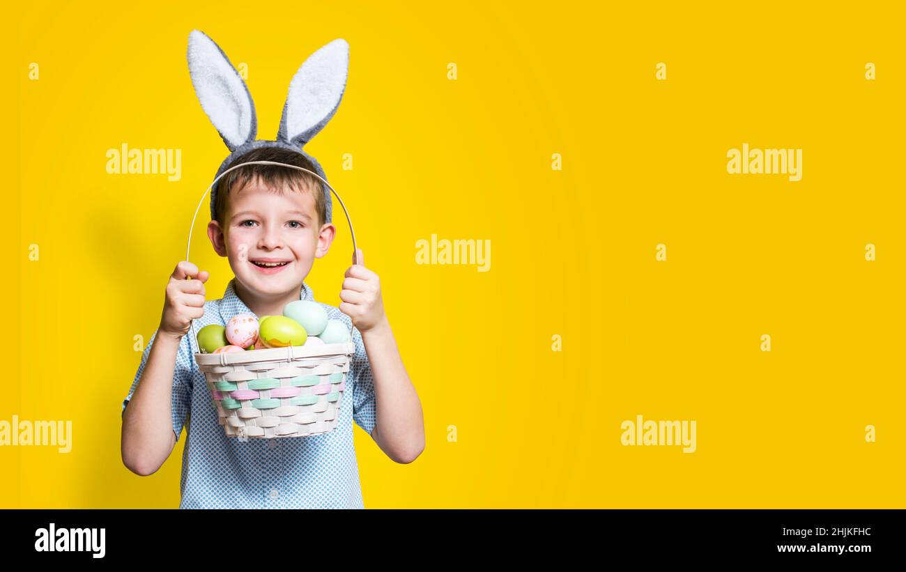 Piccolo ragazzo carino con un cestino di Pasqua in mano e coniglietto orecchie sulla testa su sfondo giallo. Sfondo di Pasqua con luogo per l'inserimento del testo. Foto Stock