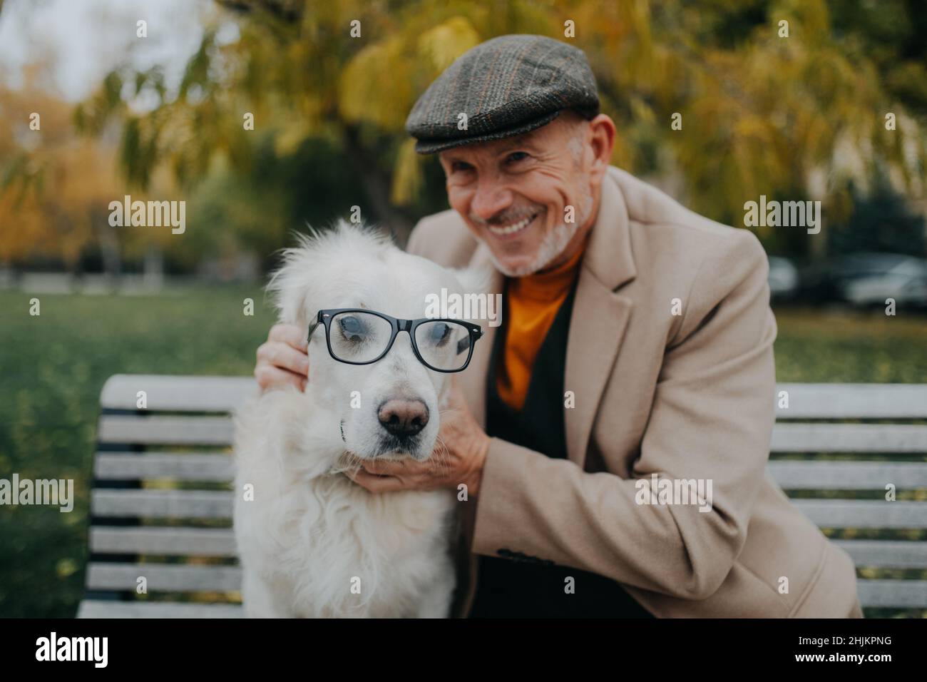 Uomo anziano felice guardando la macchina fotografica e abbracciando il suo cane indossando occhiali su panca all'aperto in città. Foto Stock