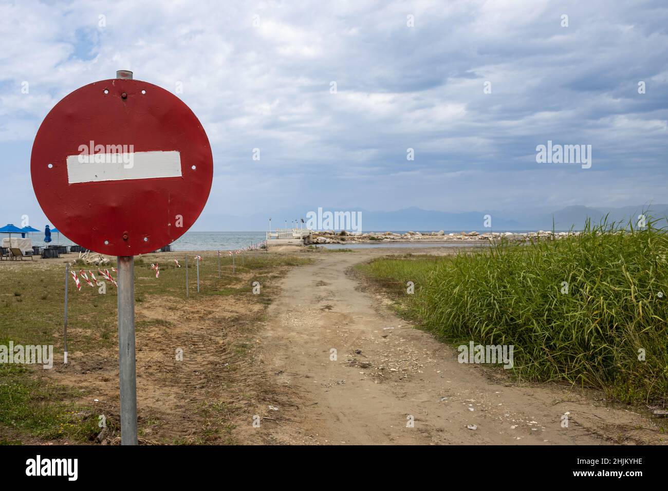 In primo piano, il simbolo di traffico meno recente non è presente. Riva del mare dello Ionio con una strada sabbiosa e verde erba di un campo. Cielo nuvoloso di primavera. Roda, corf Foto Stock