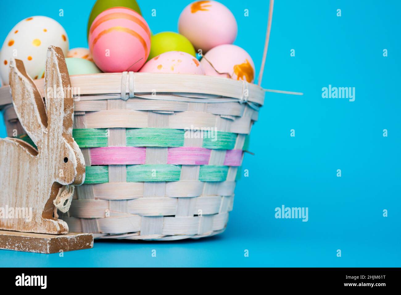 Un cestino di uova di Pasqua con coniglietto giocattolo su sfondo blu. Cestino festivo con uova colorate dipinte e coniglio di legno giocattolo. Composizione pasquale. Foto Stock
