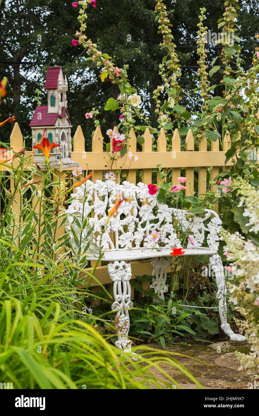 Bianco dipinto in ghisa vittoriano stile panca seduta contro beige picket recinto dipinto e delimitato da Papaver rosso - Poppy, Alcea rosea - Hollyhock. Foto Stock