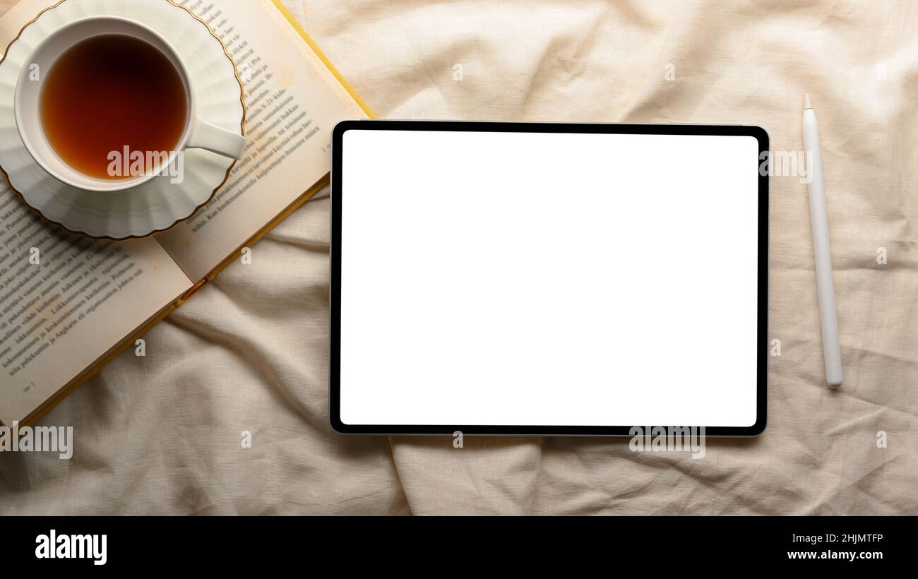 Spazio di lavoro minimale e accogliente con tazza da tè, libro di romanzo e tablet digitale schermo bianco mockup su tovaglia bianca. Vista dall'alto, disposizione piatta Foto Stock