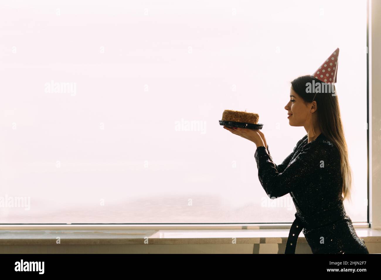 bella donna caucasica che tiene una torta guardando una candela, in piedi alla finestra dell'ufficio, sfondo chiaro, vista del profilo di una ragazza Foto Stock