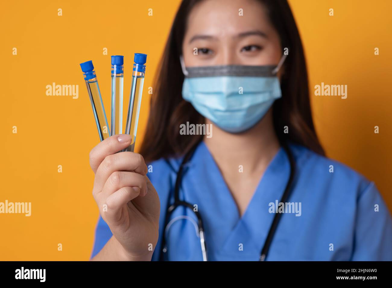 Foto studio con sfondo arancione e focalizzazione sulle provette campione nelle mani di un medico con maschera Foto Stock