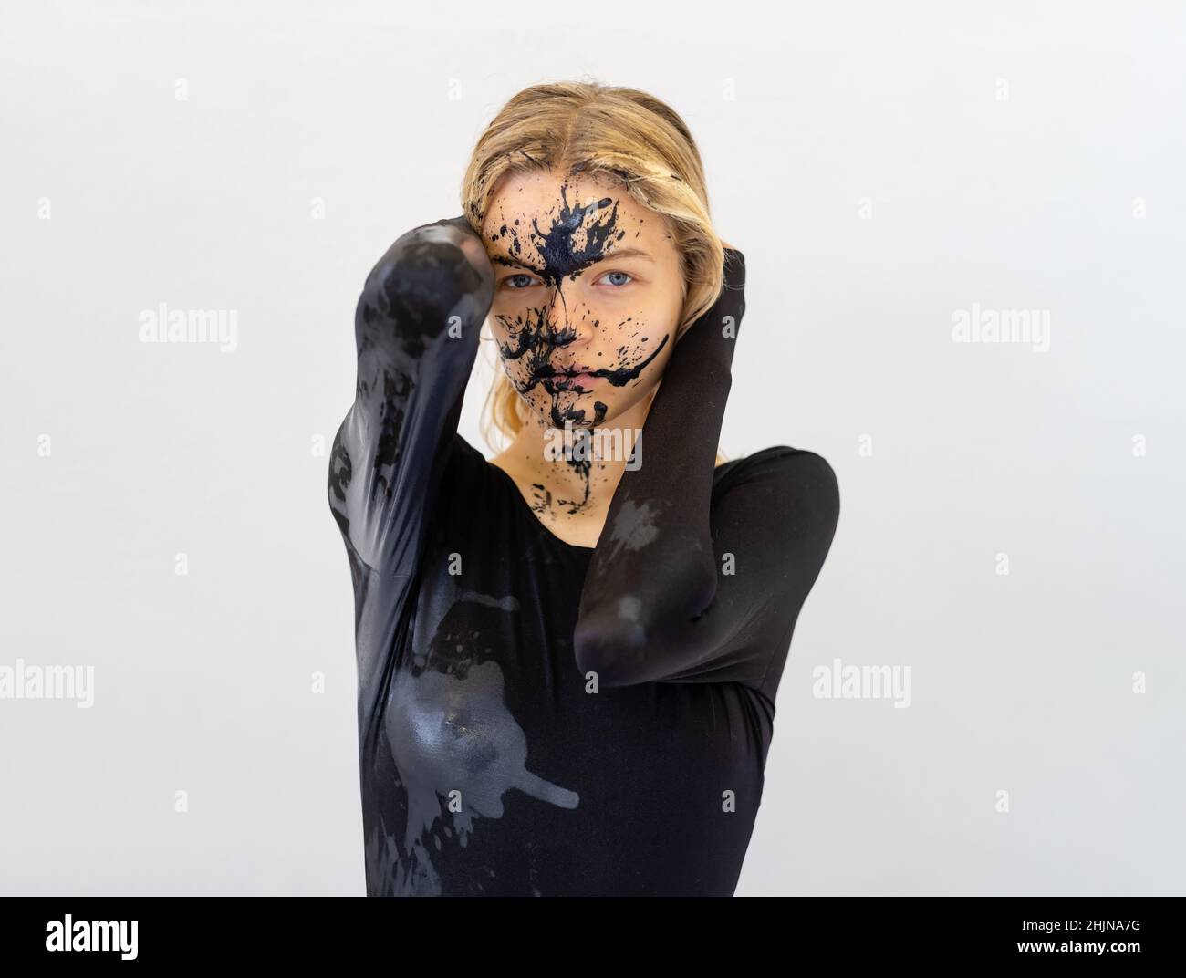 ritratto d'arte fiducioso della giovane donna bionda dipinta sexy in tuta nera con macchie di vernice nera sul viso Foto Stock
