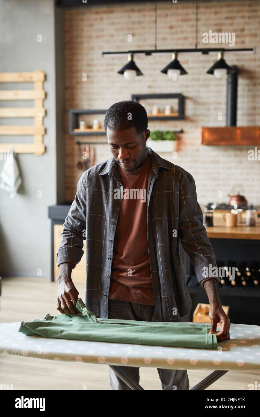 Ritratto verticale di giovane uomo afro-americano stirare vestiti in studio appartamento, stile di vita di bachelor e concetto di famiglia chores Foto Stock