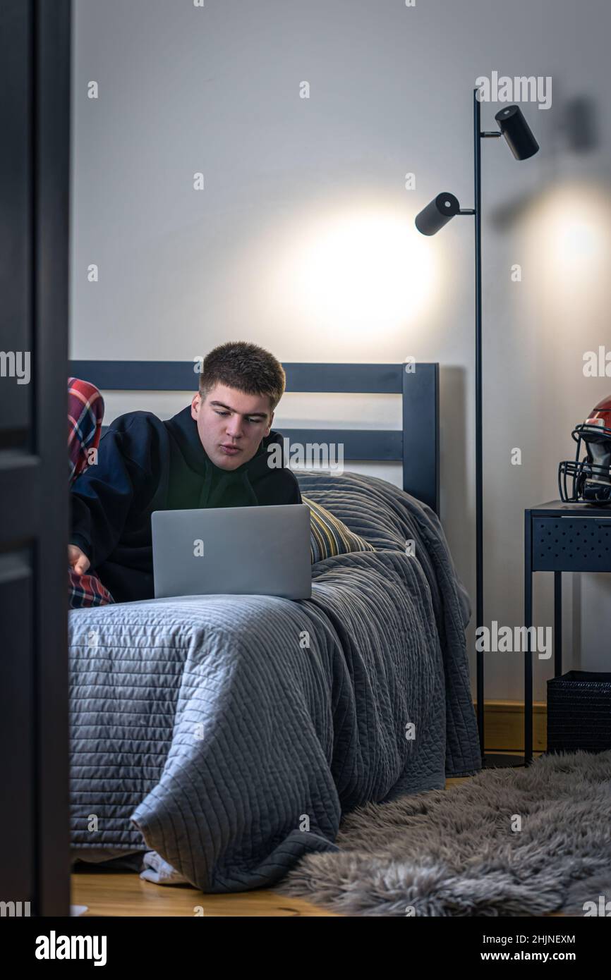 Un ragazzo si siede in una stanza su un letto e usa un computer portatile. Foto Stock
