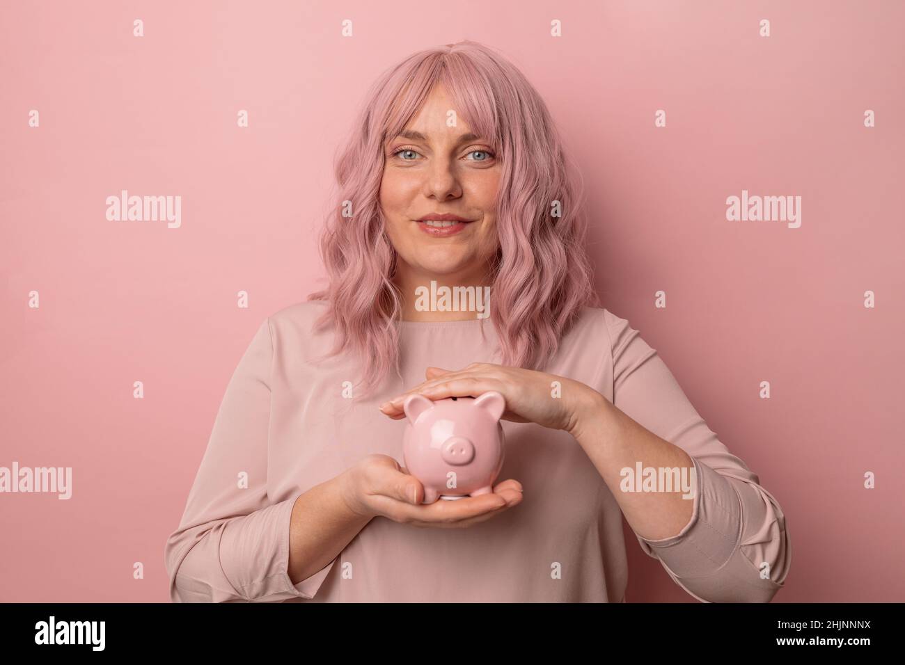 Felice positivo giovane bella attraente donna di affari sorridente tenendo un piggybank per l'investimento su sfondo rosa chiaro, copyspace Foto Stock